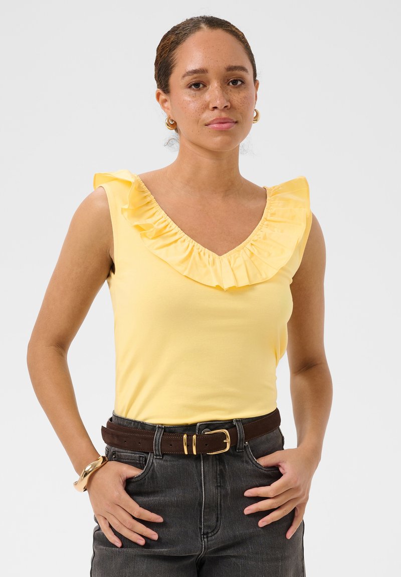 Femme portant un haut jaune sans manches avec un col à volants, un jean foncé taille haute, une ceinture marron, des boucles d'oreilles créoles dorées et un bracelet, posant les mains dans les poches.