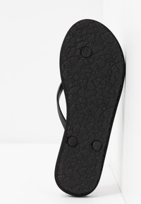 Svart flip-flop såle med et teksturert blomstermønster, flat design, to sirkulære knappe-detaljer og en minimalistisk stropp i svart.