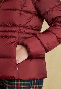 Burgundy puffer jacket with a quilted texture, featuring a zippered pocket and stitched details near the hem and sleeve.