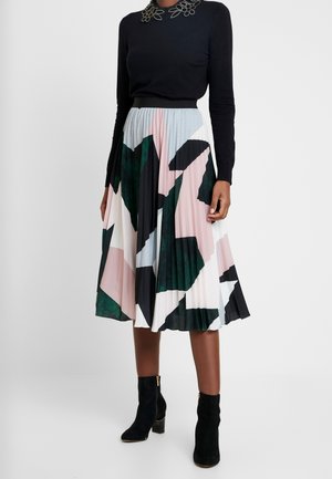 Woman wearing a black long-sleeve top with floral neckline detail, a pleated midi skirt with abstract pastel and dark color blocks, and black ankle boots.