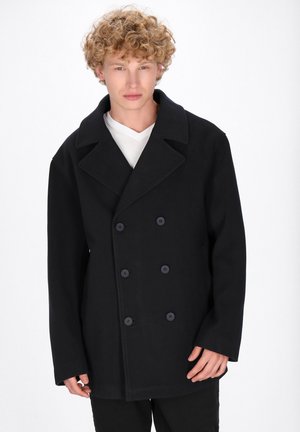 Sort uld peacoat med dobbeltbrystet design, store revers og to frontknapper. Glat tekstur, løs pasform, brugt over en hvid skjorte.
