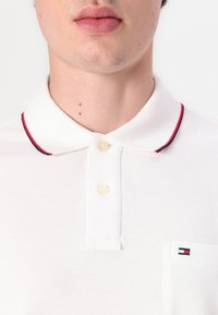 Balta polo krekls ar struktūrētu audumu, divu pogu apdare, apkakle ar sarkaniem un jūras zilas akcentiem, un neliela zīmola kabata.