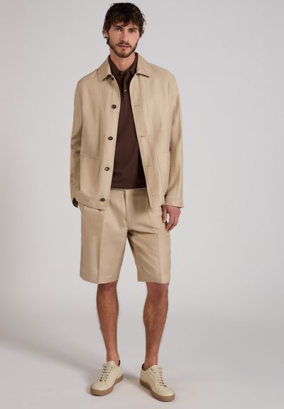 Mann trägt beigefarbenen Knopfjacke, dazu passende kniehohe Shorts, dunkelbraunes Hemd und beigefarbene Turnschuhe, steht vor einfarbigem Hintergrund.