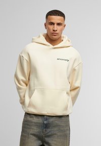 Un hoodie crème en tissu doux avec une poche avant et un logo "anomaly" vert sur la poitrine gauche. Coupe décontractée avec une capuche doublée.