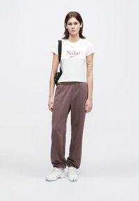 Νεαρή γυναίκα που φοράει λευκό μπλουζάκι Nike, καφέ παντελόνα, λευκά αθλητικά παπούτσια και μαύρη τσάντα σώματος, στέκε ται μπροστά σε απλό λευκό φόντο.