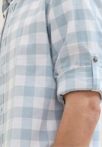 Chemise à manches courtes à carreaux bleu clair et blanc avec un poignet boutonné, présentant une texture de tissu douce et des détails de couture visibles.