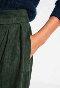 Falda de ante suave de color verde oscuro con pliegues, que cuenta con bolsillos laterales. Una mano se introduce en uno de los bolsillos, destacando el diseño de la falda.