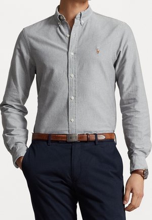 Hombre con camisa de botones gris claro con pequeño logo bordado, pantalones azul marino oscuro, cinturón de cuero marrón y reloj de pulsera, con las manos en los bolsillos.