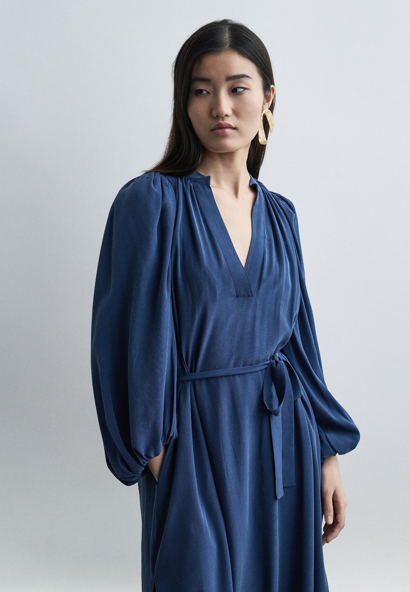 Γυναίκα με μακριά σκούρα μαλλιά φοράει ένα μπλε φόρεμα με ζώνη, φουσκωτά μανίκια και μεγάλο στριφτό χρυσό κρίκο στο αυτί, κοιτάζοντας πλάγια.