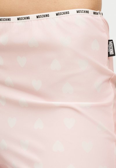 Ροζ ανοιχτό ύφασμα με διακριτικά μοτίβα καρδιών, λευκή ζώνη μέσης με επαναλαμβανόμενο κείμενο "Moschino" και μια μαύρη ετικέτα Moschino Active στο πλάι.