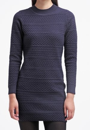Femme portant une robe en maille bleu marine ajustée à manches longues avec des motifs en torsades et en tissage panier, associée à des collants noirs.