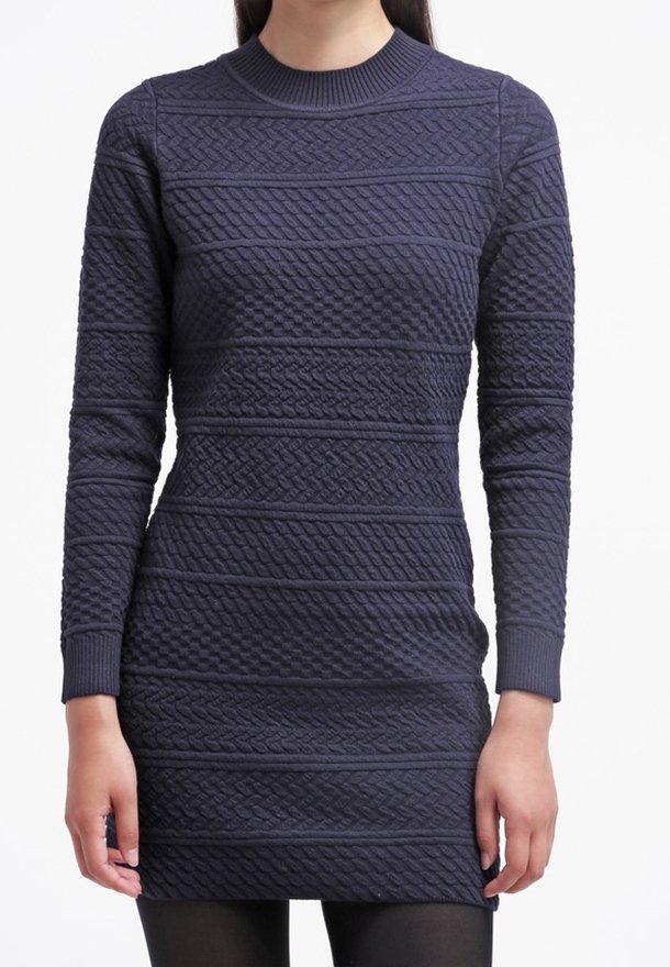 Femme portant une robe en maille bleu marine ajustée à manches longues avec des motifs en torsades et en tissage panier, associée à des collants noirs.