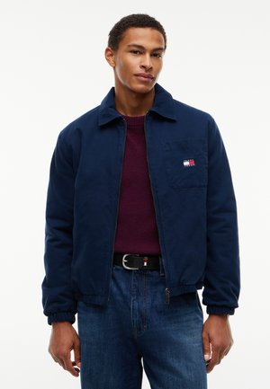 Man draagt een marineblauw jack met een logo op de voorzak, een bordeauxrode trui, blauwe spijkerbroek en een zwarte riem, staand tegen een effen witte achtergrond.