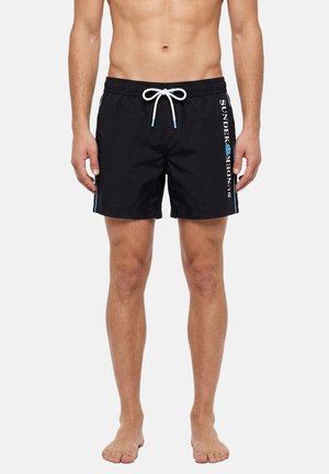 Uomo a torso nudo che indossa pantaloncini da bagno neri con coulisse bianca e logo verticale "SUNDEK" sulla gamba destra, in piedi a piedi nudi su uno sfondo neutro.