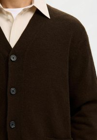 Brauner Strickcardigan mit V-Ausschnitt, langen Ärmeln und vier schwarzen Knöpfen. Über einem hellgestreiften Hemd getragen. Strukturierter Stoff.