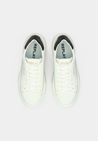 Λευκά sneakers με λείο δέρμα, στρογγυλερή μύτη, επίπεδα λευκά κορδόνια και αντίθετη μαύρη λεπτομέρεια στο τακούνι. Η επωνυμία "REPLAY" στο γλώσσα.