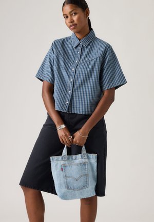 Jonge vrouw die een blauw geruit overhemd met korte mouwen en een zwarte rok draagt, met in haar hand een lichtblauwe denim draagtas met een voorvak.