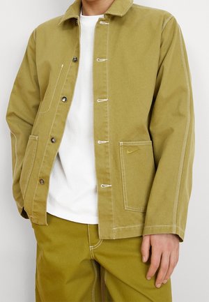 Man draagt een olijfgroene jas met witte stiksels, wit T-shirt en bijpassende olijfgroene broek, staat met één hand naar beneden.