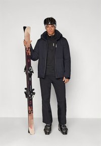 Completo da sci con giacca imbottita scura, maglia termica nera e pantaloni neri. Tiene gli sci con dettagli rossi, indossa un casco nero e occhiali da sci.