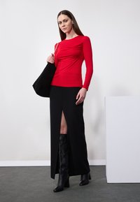 Červený top s dlhým rukávom skombinovaný s čiernou maxi sukňou s vysokým rozparkom. Modelka nesie čiernu semišovú tašku a má na sebe čierne kožené čižmy nad kolená.