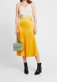 Gonna midi gialla in satin con una finitura testurizzata, abbinata a un top beige. Porta una borsa verde con un'accenno circolare dorato. Stivali neri alla caviglia.