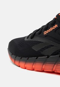Sort Reebok løbesko med mesh-overdel, sorte snørebånd og orange gradient segmenteret sål samt logo på siden.
