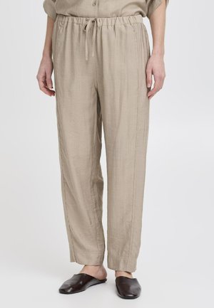 Light beige relaxed pants with an elastic waistband and drawstring; soft, textured fabric with side seams. Worn with dark slip-on shoes.