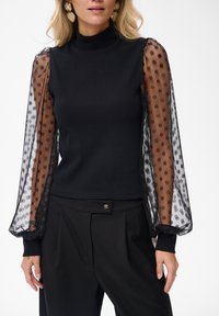 Czarny prążkowany top z wysokim dekoltem, z przezroczystymi puffami z brązowymi kropkami. W komplecie z dopasowanymi czarnymi spodniami.