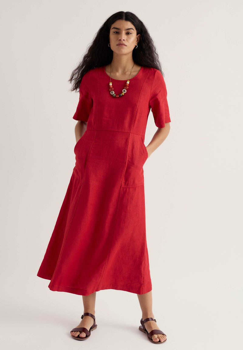 Vrouw met lang krullend haar, gekleed in een rode midi-jurk met zakken, een kralenketting en bruine sandalen, staand tegen een effen achtergrond.
