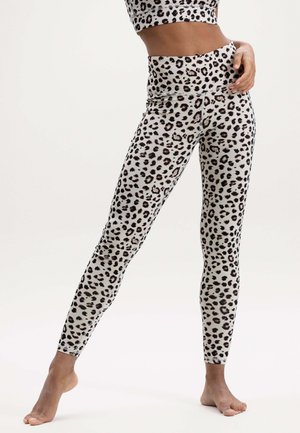 Frau trägt hoch taillierte Leggings mit Leopardenmuster und passendes kurzes Oberteil, steht barfuß mit einem Bein seitlich ausgestreckt.