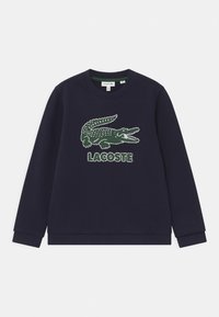Marinblå sweatshirt gjord av bomull. Har en stor grön krokodil-logotyp med prickig textur och "LACOSTE"-text nedanför. Standard rundad hals.