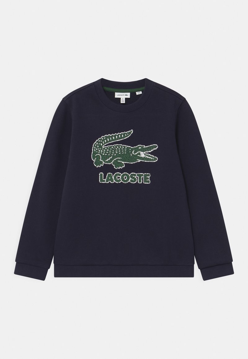 Marinblå sweatshirt gjord av bomull. Har en stor grön krokodil-logotyp med prickig textur och "LACOSTE"-text nedanför. Standard rundad hals.