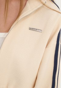 Chaqueta con cremallera en tela de textura de gofre color crema, con rayas azul marino, una placa de logo de herrajes plateados y un diseño de cuello alto.