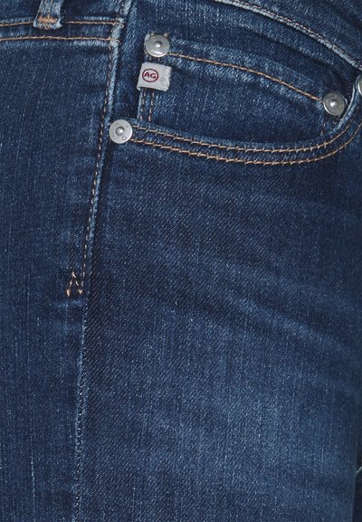 Jeans de ganga azul-escuro com acessórios prateados, costura laranja em contraste e uma pequena etiqueta com o logotipo da AG no bolso da frente.