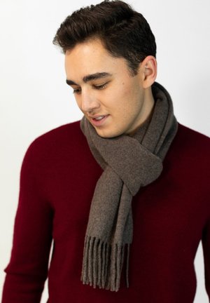 Young man wearing a burgundy sweater and a brown fringed scarf tied around his neck, looking downward with a slight smile.