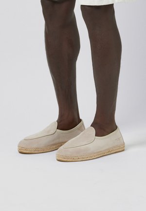 Donkere benen die beige suède instap-espadrilles met juten zolen dragen tegen een effen lichte achtergrond.