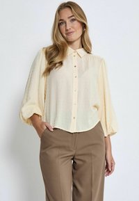 Hellgelbe Bluse mit spitzem Kragen und Knopfleiste. Puffärmel mit strukturiertem Punktemuster, kombiniert mit braunen Hosen.