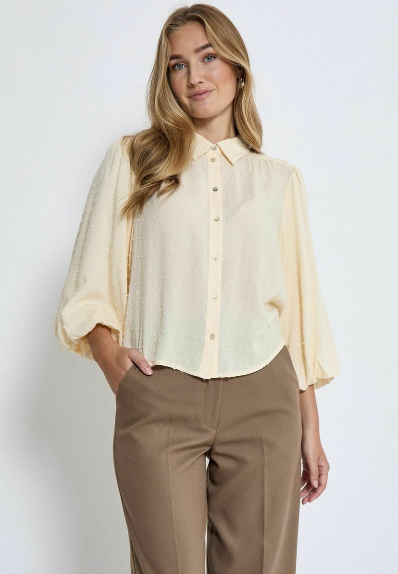 Hellgelbe Bluse mit spitzem Kragen und Knopfleiste. Puffärmel mit strukturiertem Punktemuster, kombiniert mit braunen Hosen.