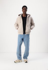 Beige hoodie met een rits aan de voorkant en zakken, gedragen over een zwarte, textuurtrui, gecombineerd met lichtblauwe, baggy jeans en beige sneakers.