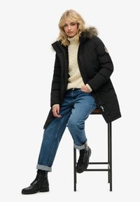 Czarna parkas z futrzanym kapturem, z zamkiem i zapięciem na napy oraz kieszeniami po bokach. Nosi dżinsy z mankietami w kolorze niebieskim oraz sweter w kremowym kolorze z grubym splotem.