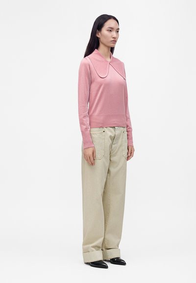Femme portant un haut rose à manches longues avec un grand col et un pantalon beige à jambes larges, debout devant un fond blanc uni.