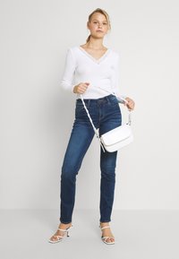 Weißes Langarmoberteil mit Spitzensaum, blaue enge Jeans, weiße strukturierten Umhängetasche und weiße Riemchensandalen. Neutraler Hintergrund.