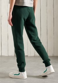 Persoon gekleed in donkergroene sportbroek en witte sneakers met groene streep, staande op een grijze vloer tegen een witte houten paneelwand.