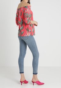 Top rojo de un solo hombro con estampado de hojas tropicales, combinado con jeans ajustados azul claro y mules de tacón alto rosas. Tejido suave y diseño ajustado.