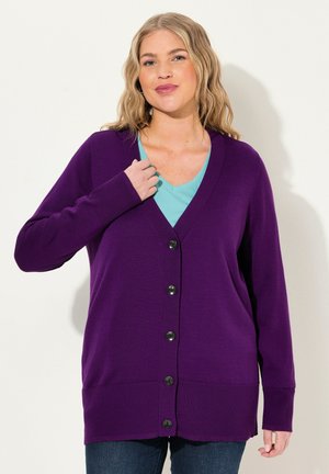 Lila Strickjacke mit langen Ärmeln, V-Ausschnitt, fünf schwarzen Knöpfen und gerippten Bündchen/Saum, über einem hellblauen Oberteil getragen.