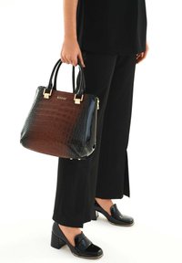 Sac à main en cuir embossé crocodile marron et noir avec quincaillerie dorée, poignées doubles et fermeture éclair. Porté avec un pantalon et des chaussures noirs.