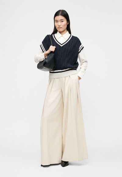 Femme debout portant un pull sans manches en maille torsadée bleu marine par-dessus une chemise blanche, un pantalon large plissé crème, des chaussures noires, et tenant un sac à main noir.