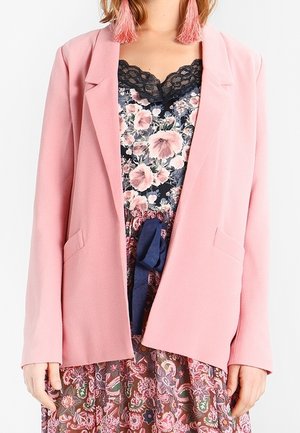 Femme portant un blazer rose sur un haut fleuri orné de dentelle et une jupe à motifs avec une ceinture en tissu bleu marine.