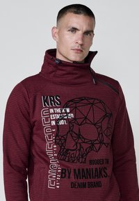Suéter rojo con estampado, cuello alto, gráfico de calavera geométrica negra y texto blanco. Presenta un cuello con cremallera y material texturizado.