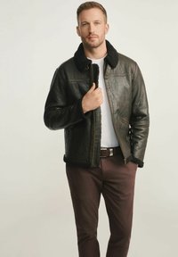 Brązowa skórzana kurtka z czarnym kołnierzem z owczej wełny, z zamek błyskawicznym i teksturowaną powierzchnią. Nosi ją nad białą koszulą i brązowymi spodniami.